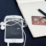 A white smartphone with earbuds, credit card, and pen on a notepad, symbolizing tech and finance.